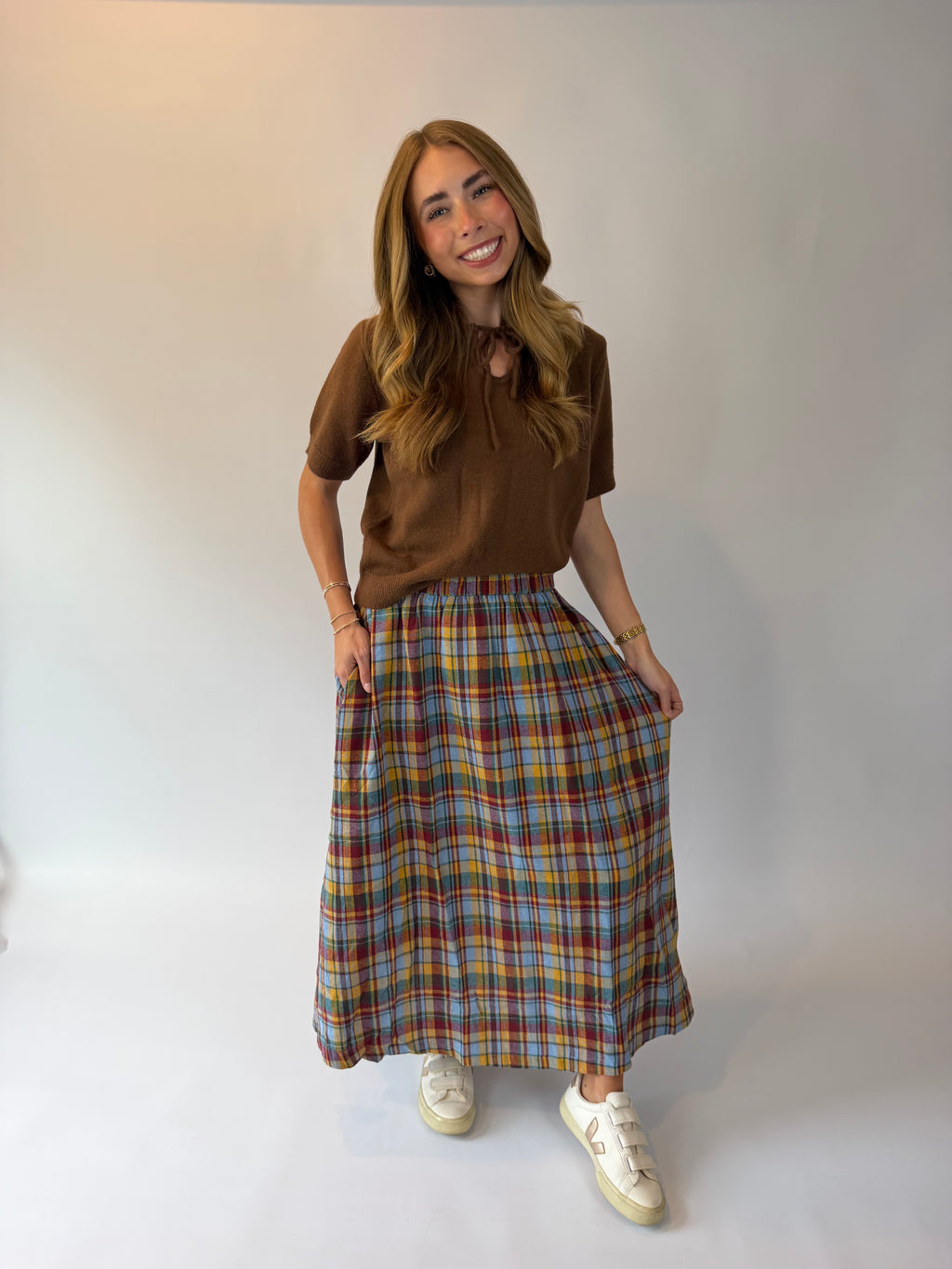 A woman modeling a brown short-sleeve top and a long multicolor plaid skirt with white sneakers, standing against a plain light gray studio background.