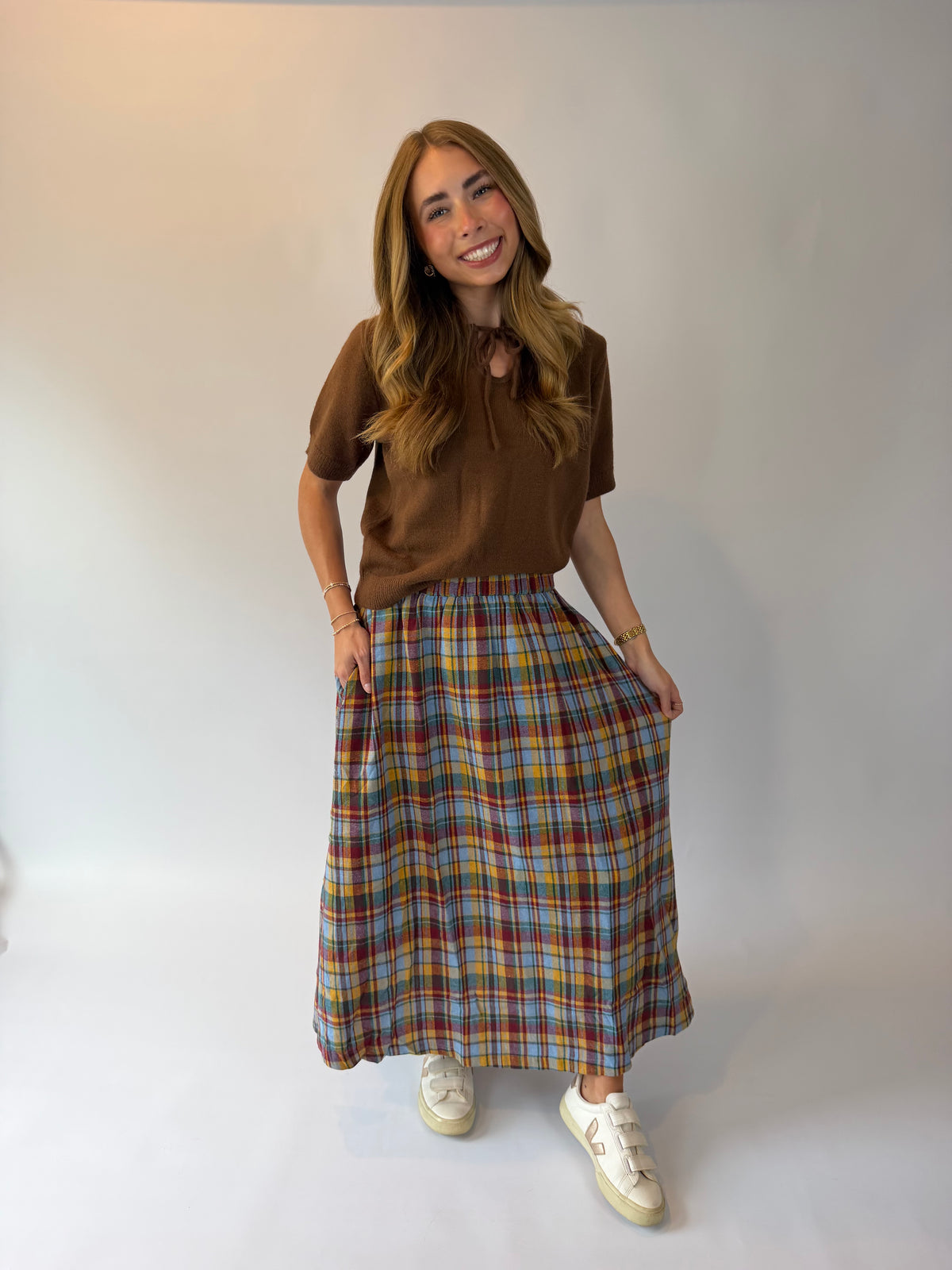 A woman modeling a brown short-sleeve top and a long multicolor plaid skirt with white sneakers, standing against a plain light gray studio background.