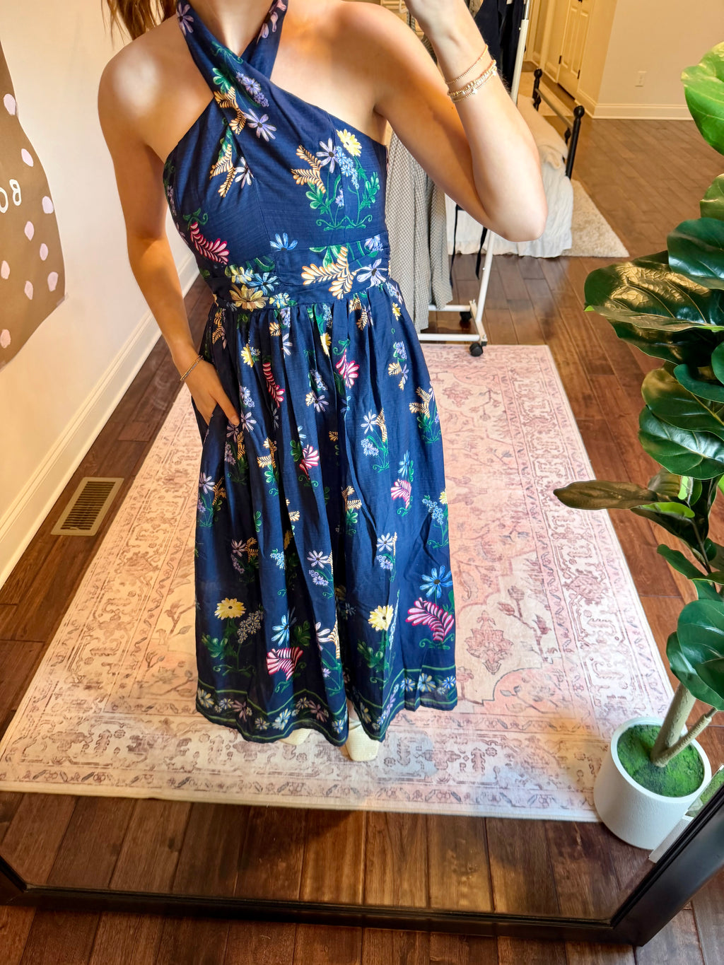 Woman taking a mirror selfie wearing a blue floral dress  in a room with a plant and decorative wall art.