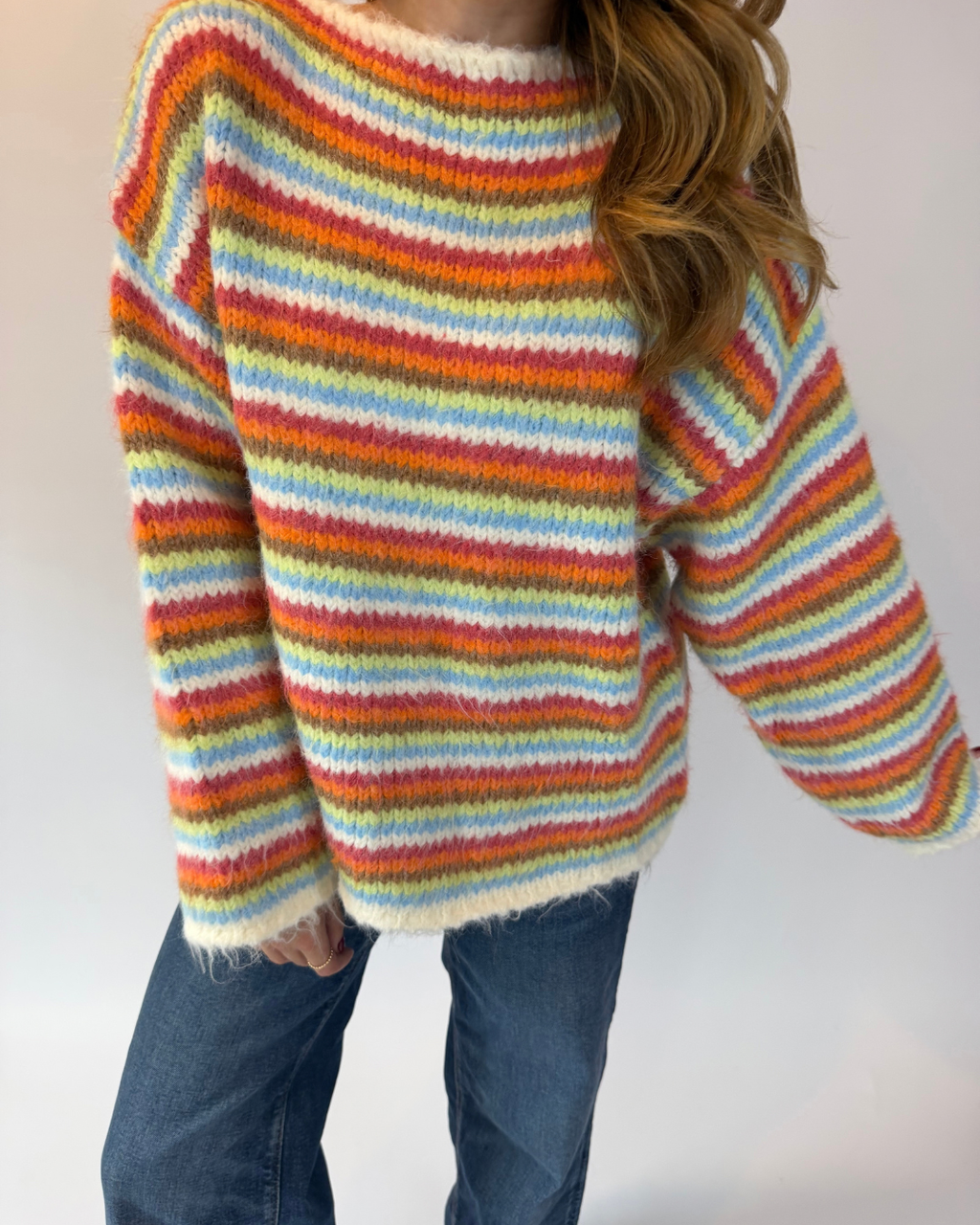 A person wearing a fuzzy striped knit sweater with horizontal bands of orange, yellow, green, blue, and cream, paired with blue jeans against a plain light background.