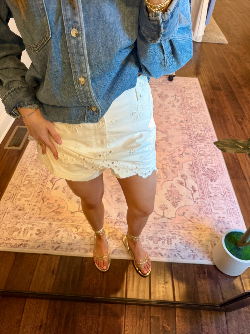 Person taking a mirror selfie wearing a denim jacket and white lace skirt in a room with a rug and plant.