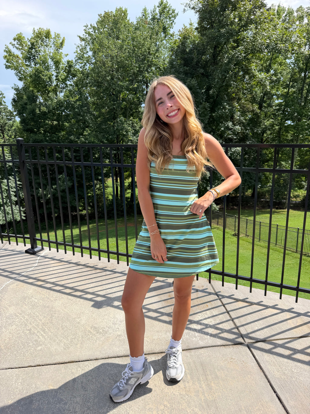A woman stands outdoors on a concrete patio wearing a sleeveless, short A-line mini dress with horizontal stripes in light blue, green, and white, paired with white socks and gray sneakers. A black metal railing and green trees are visible in the background.