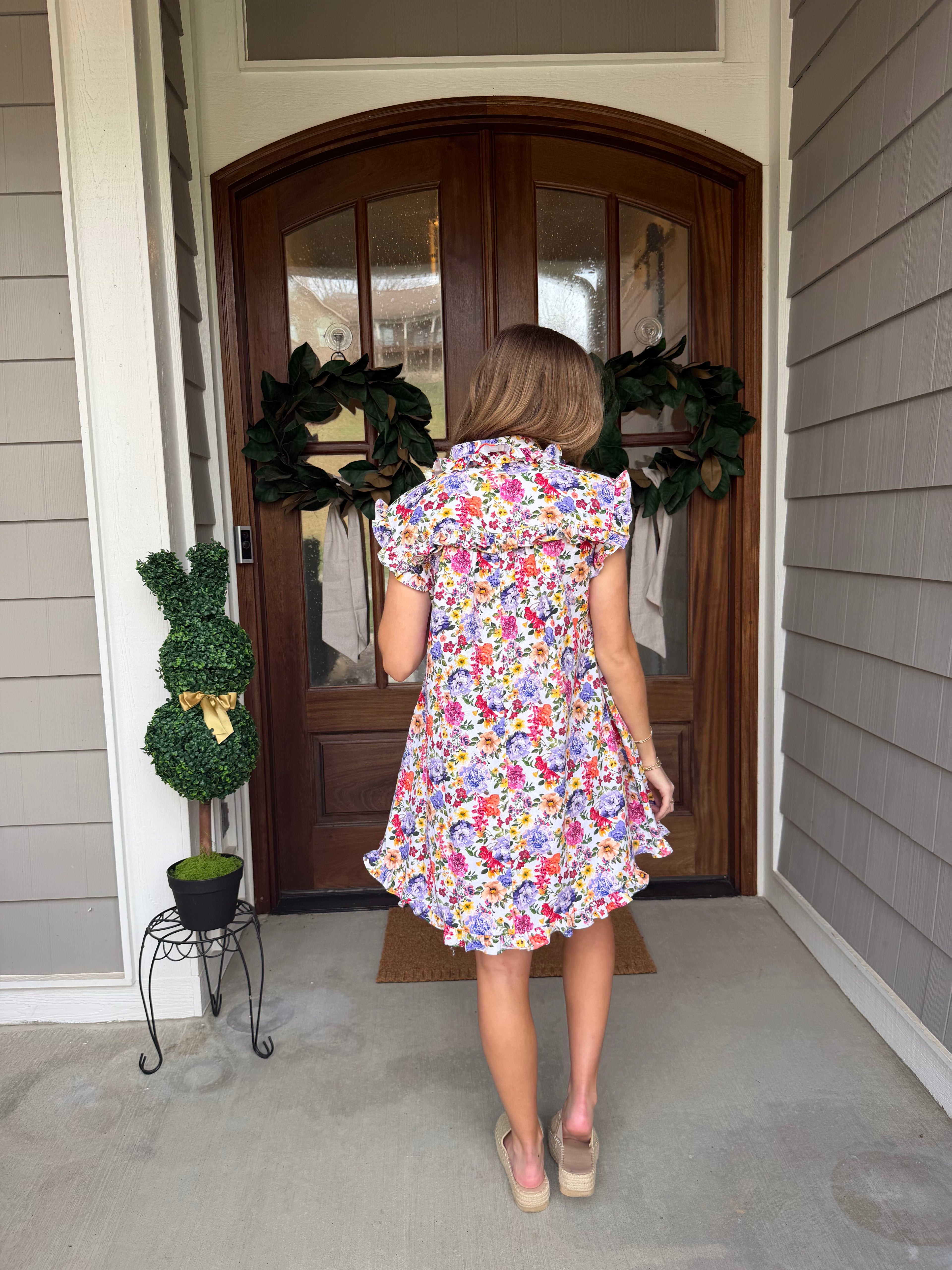 Woman in a floral dress standing in front of a wooden door with plants around.