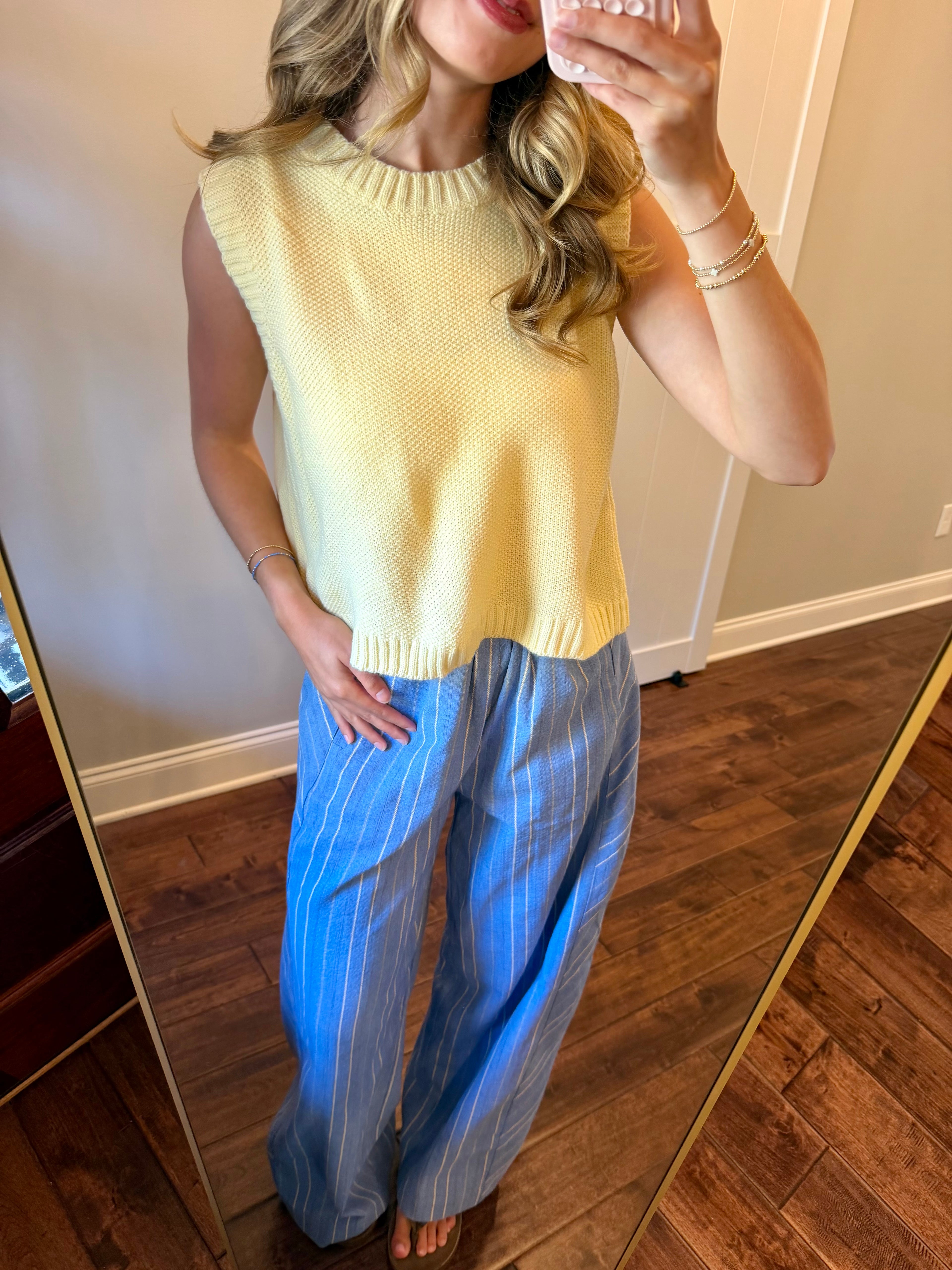 Woman wearing a yellow sleeveless top and blue striped pants, standing in front of a mirror.