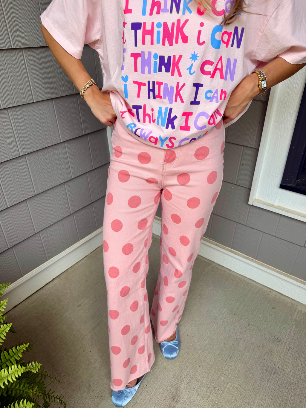 A person wearing a light pink graphic T-shirt and matching pink pants with darker pink polka dots, standing outdoors against gray house siding.