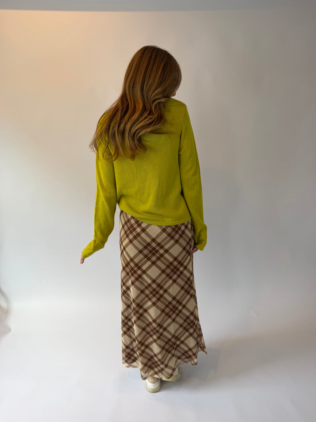 A woman is shown from behind wearing a bright yellow-green knit sweater and a brown and beige plaid maxi skirt, standing against a plain white studio background.