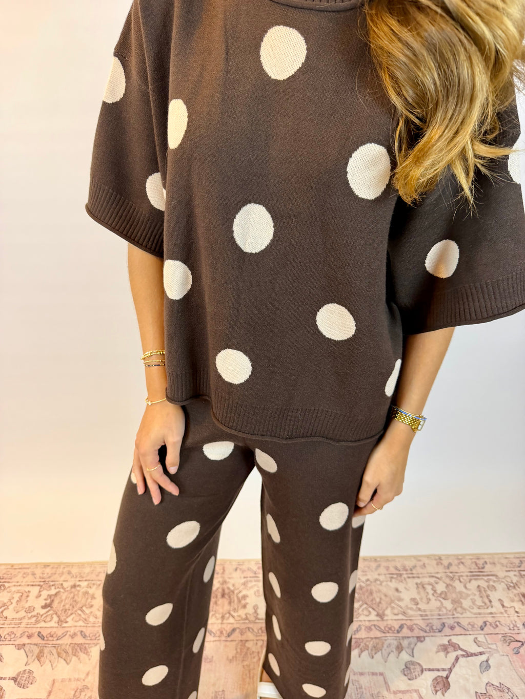 A woman wearing a matching brown knit lounge set with large white polka dots, featuring a short-sleeve top and wide-leg pants, standing against a light wall.