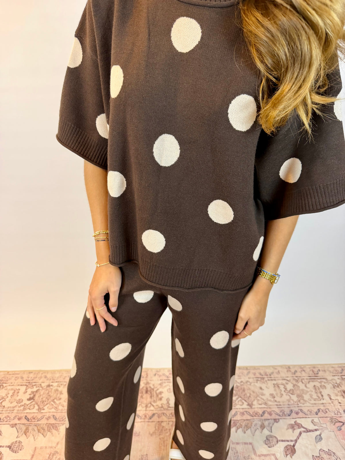 A woman wearing a matching brown knit lounge set with large white polka dots, featuring a short-sleeve top and wide-leg pants, standing against a light wall.
