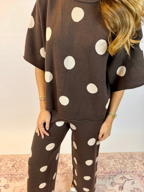 A woman wearing a matching brown knit lounge set with large white polka dots, featuring a short-sleeve top and wide-leg pants, standing against a light wall.