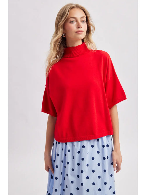 A woman wearing a bright red short-sleeve mock neck knit top and a light blue skirt with dark blue polka dots, photographed against a light gray studio background.