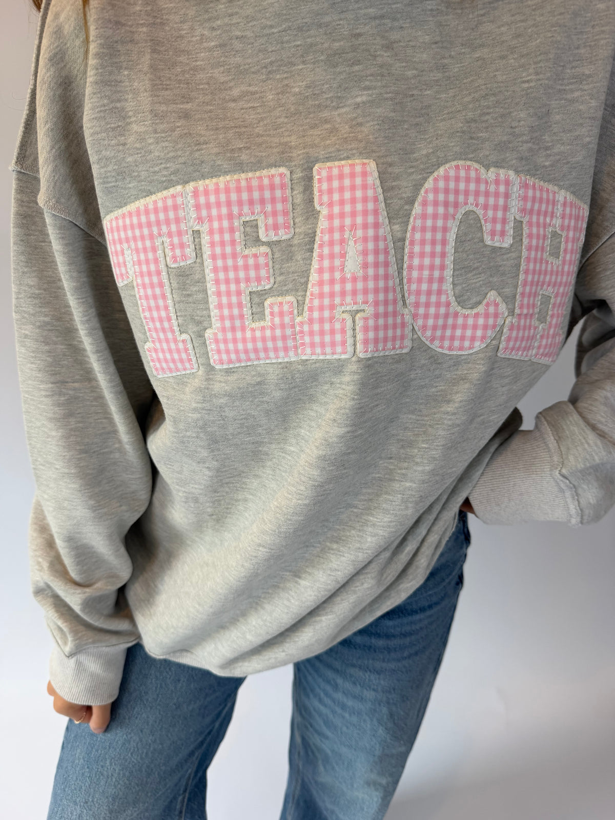 A person wearing a heather gray sweatshirt with the word 'TEACH' across the chest in pink and white gingham letters, paired with blue jeans.