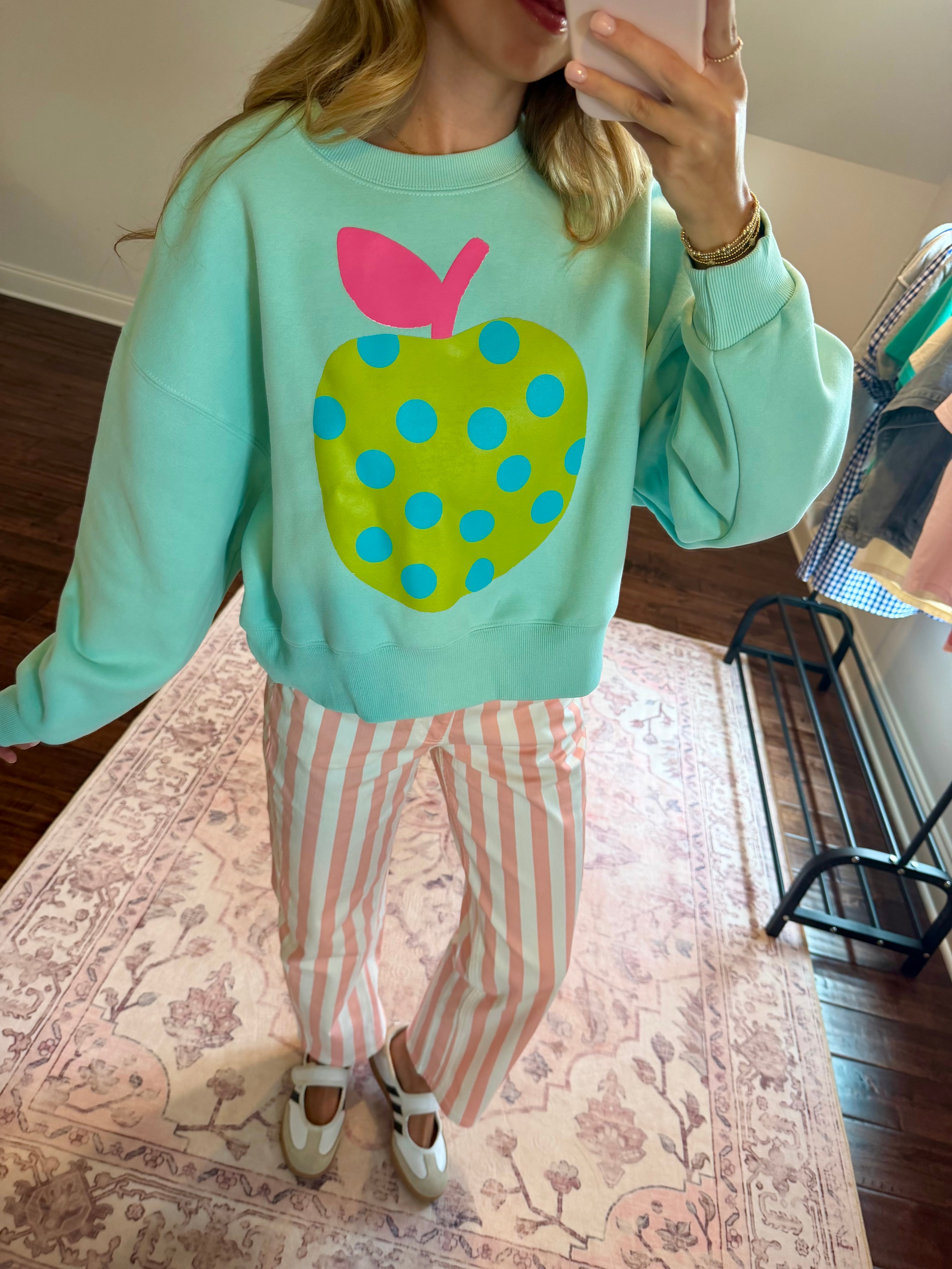 A woman takes a mirror selfie wearing a mint green sweatshirt with a large green apple graphic featuring blue polka dots and a pink stem, paired with pink and white striped pants.