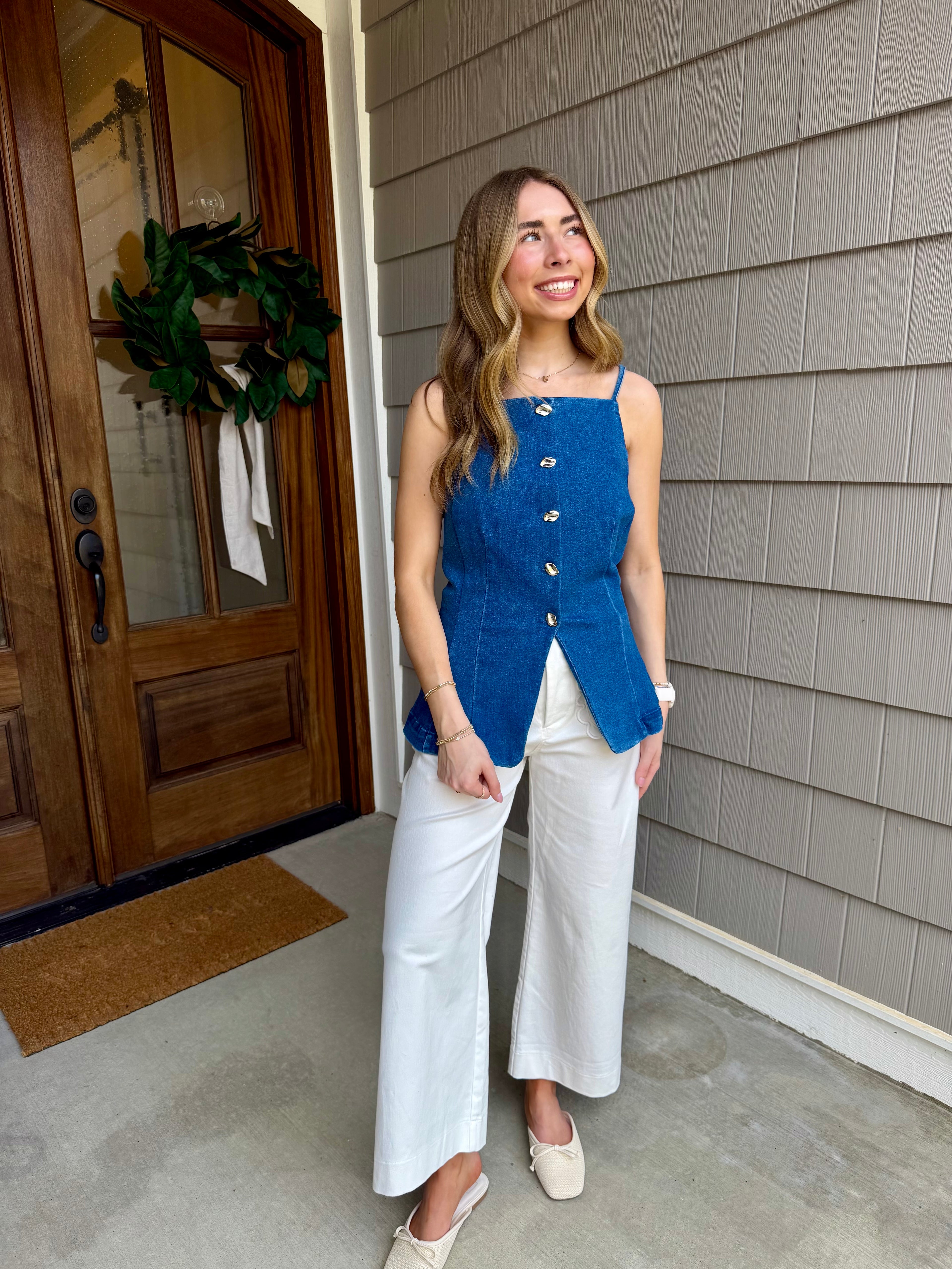 Woman in a blue vest and white pants standing on a porch.