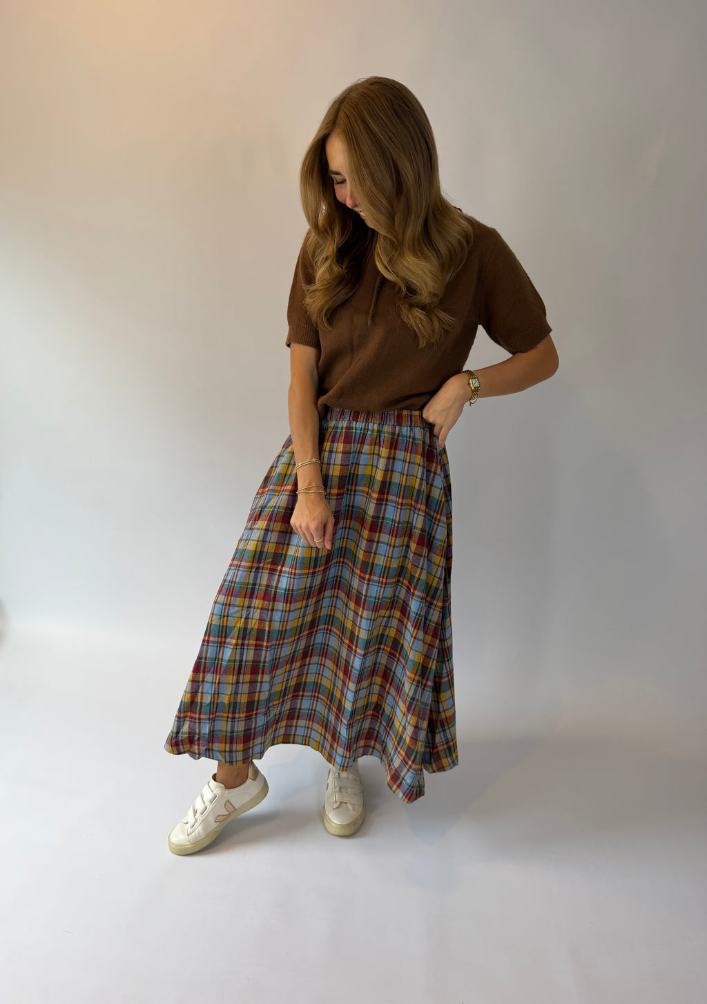 A woman modeling a brown short-sleeve polo shirt and a long multicolor plaid skirt with white sneakers, standing against a plain light gray studio background.