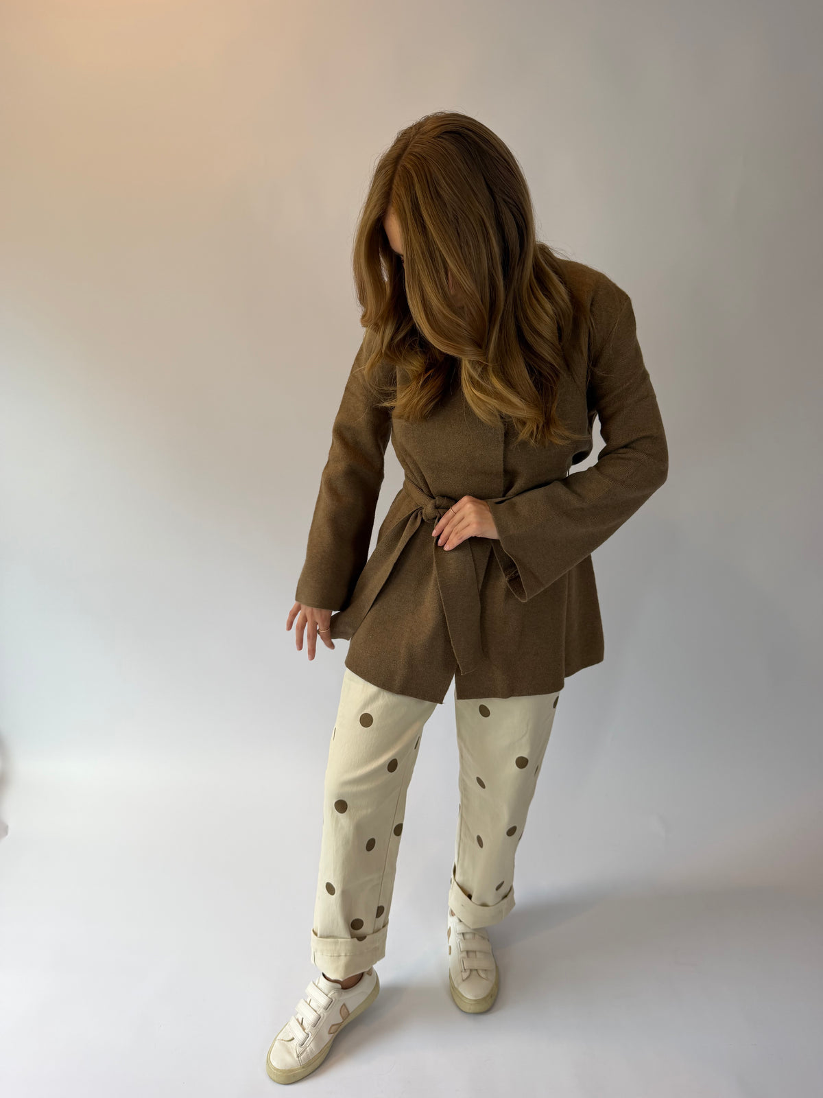 A woman modeling a brown belted coat over cream pants with brown polka dots and white sneakers, standing against a plain light gray studio background.