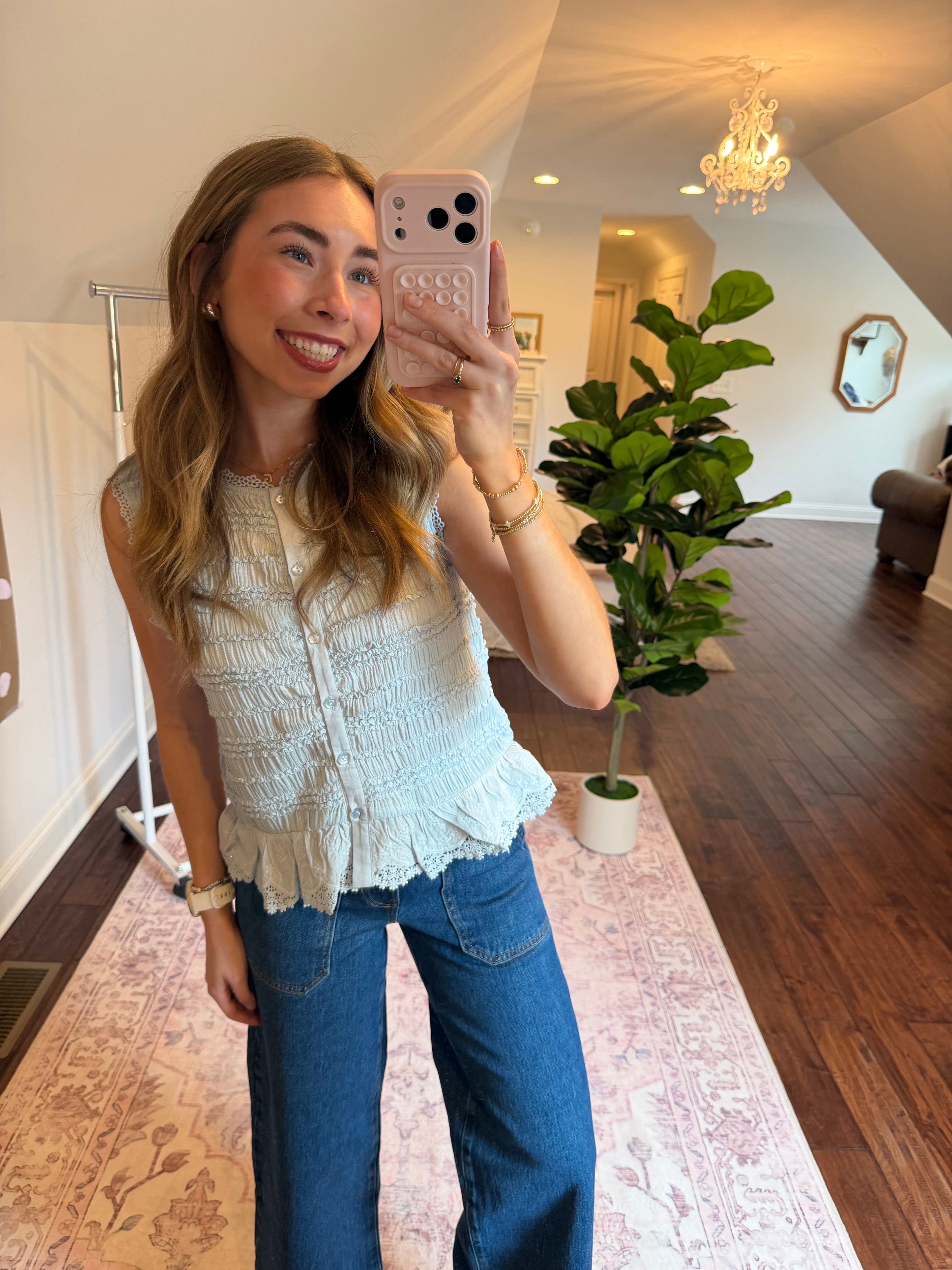 Woman taking a selfie in a room with a plant and decorative items. Wearing a baby blue tank top.