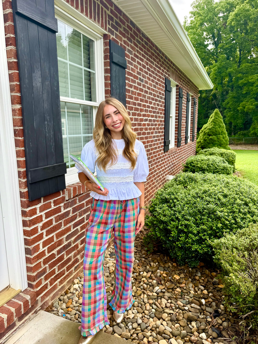 A woman stands outdoors in front of a brick house, wearing a light blue ruffled short-sleeve top and colorful plaid flare pants, holding a notebook.