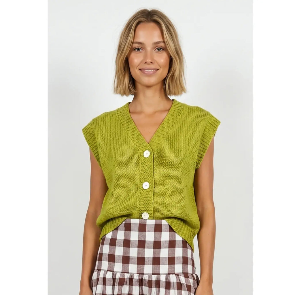A woman wearing a lime green ribbed knit sleeveless cardigan with white buttons, paired with a brown and white gingham skirt, against a light gray studio background.
