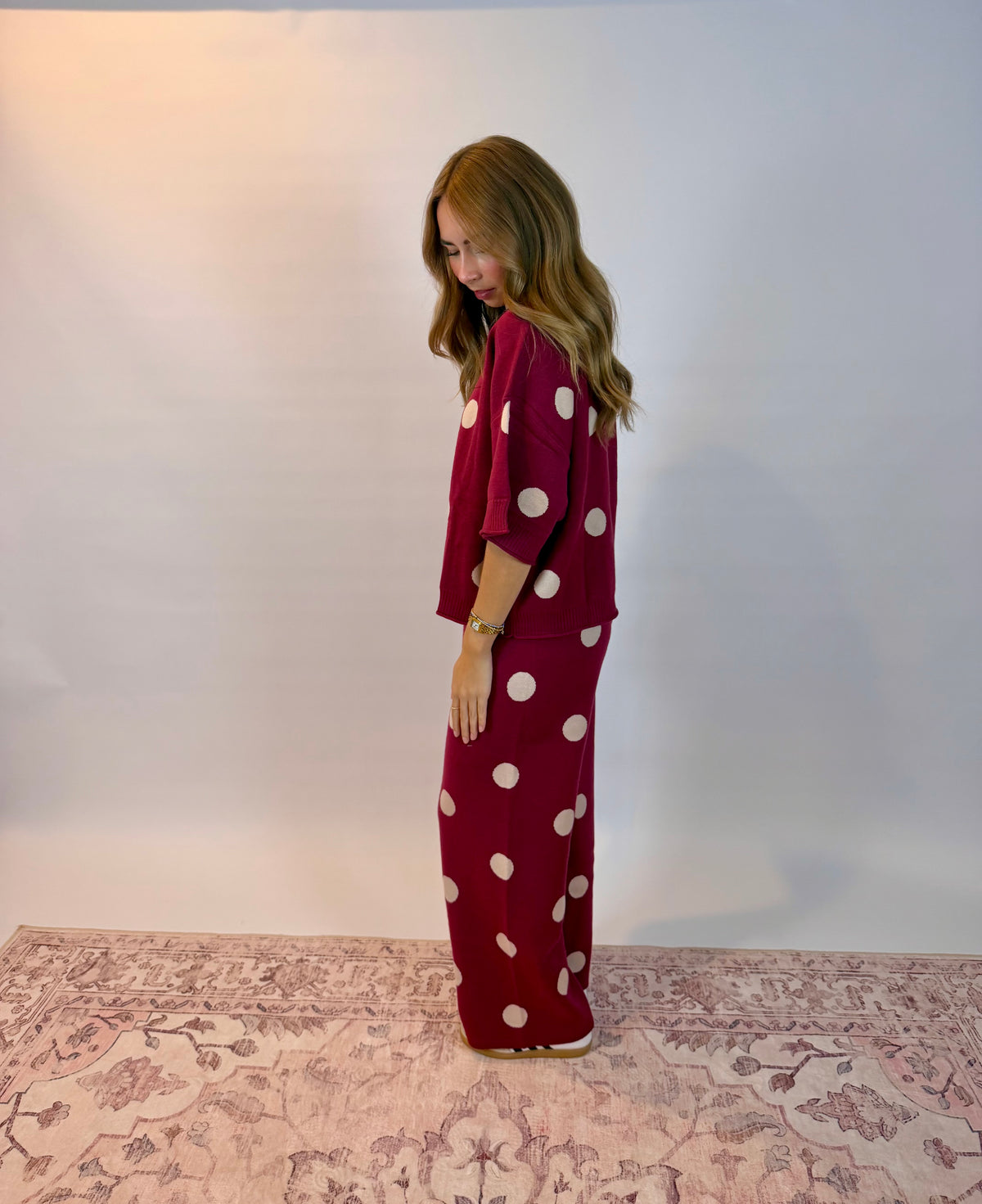 A woman stands in profile wearing a matching two-piece lounge set in burgundy with large white polka dots, standing on a patterned rug against a plain light gray studio backdrop.