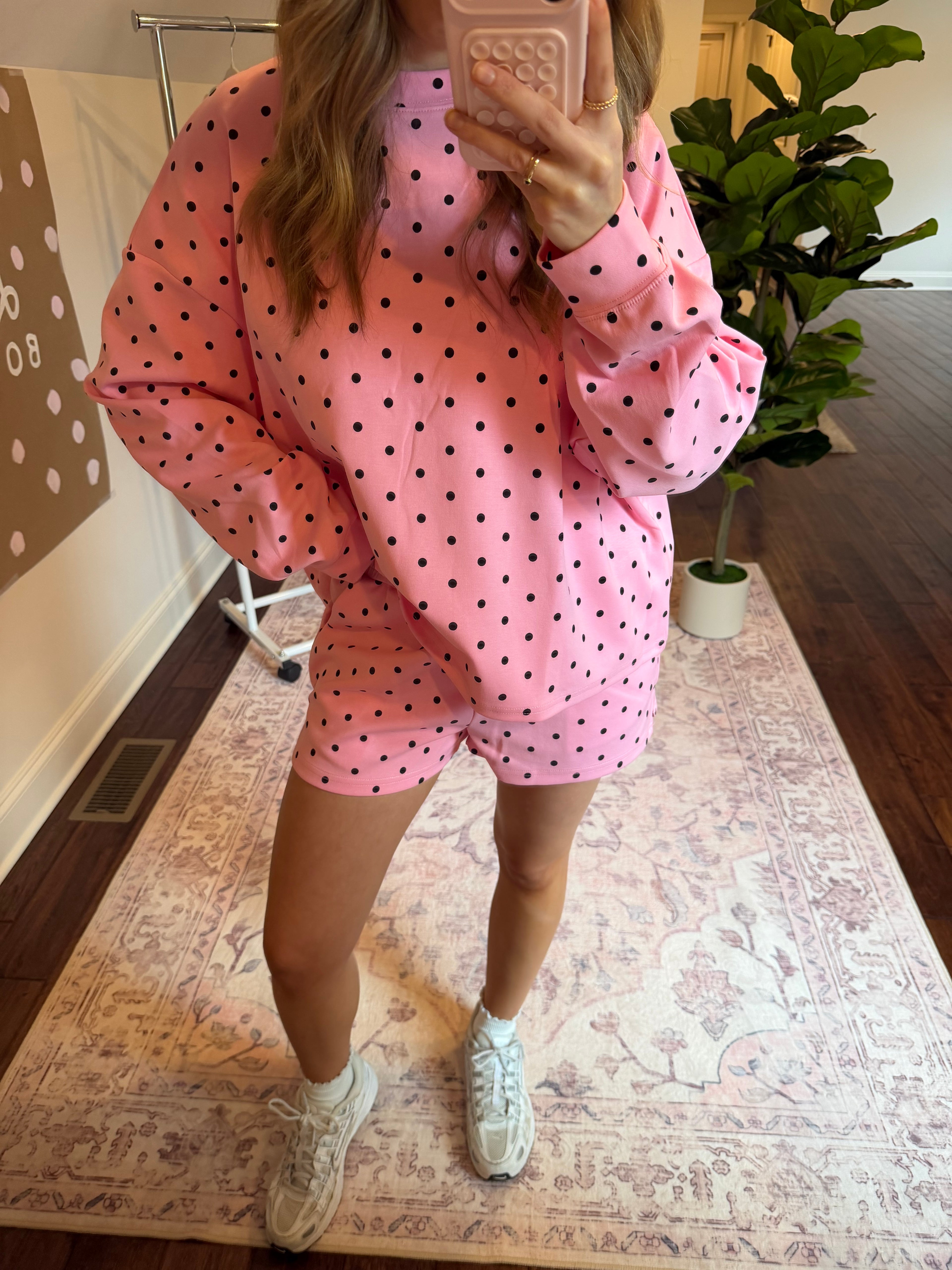Woman in a pink polka dot matching set standing in a room with a plant and patterned wall.