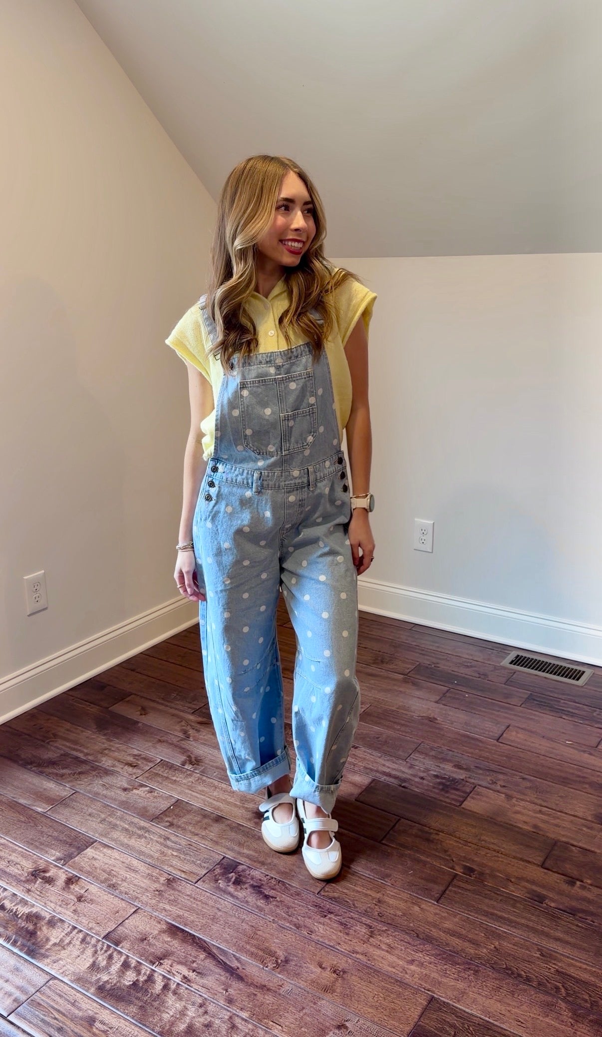 Woman wearing a yellow top and light blue overalls standing in a room with wooden flooring.