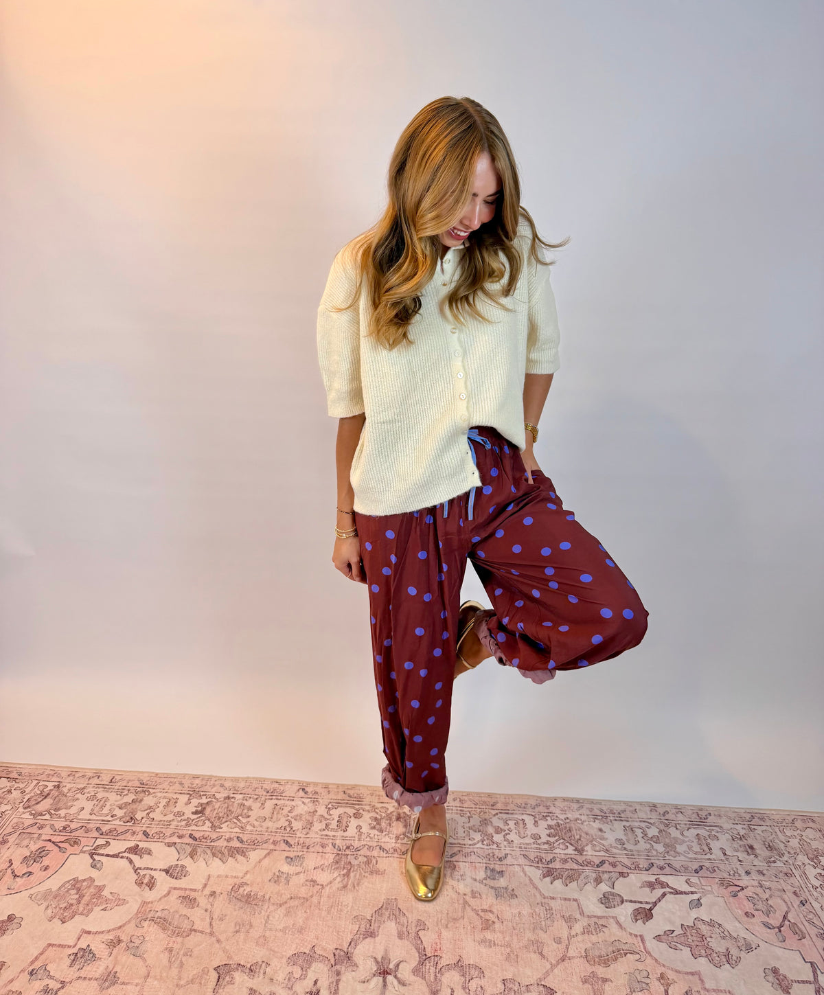 A woman posing in a cream-colored short-sleeve button-up shirt and burgundy pants with blue polka dots, standing on a patterned rug against a plain studio backdrop.
