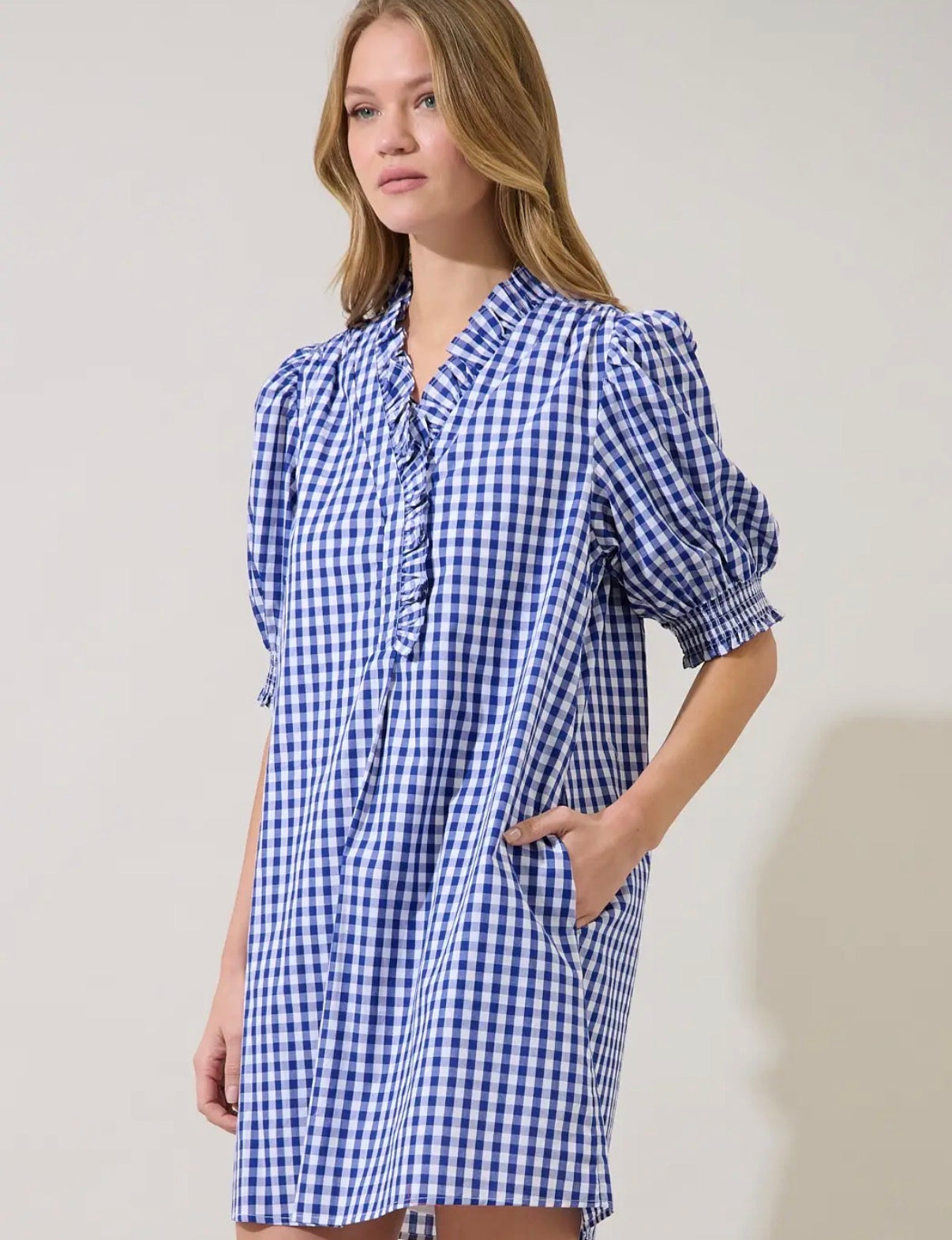 A woman wearing a blue and white gingham mini dress with puff sleeves, a ruffled tie neckline, and side pockets, photographed against a plain light beige studio background.