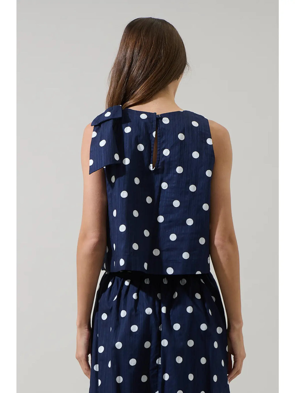 A woman shown from the back wearing a navy blue sleeveless dress with a white polka dot pattern and a large bow detail on the shoulder.