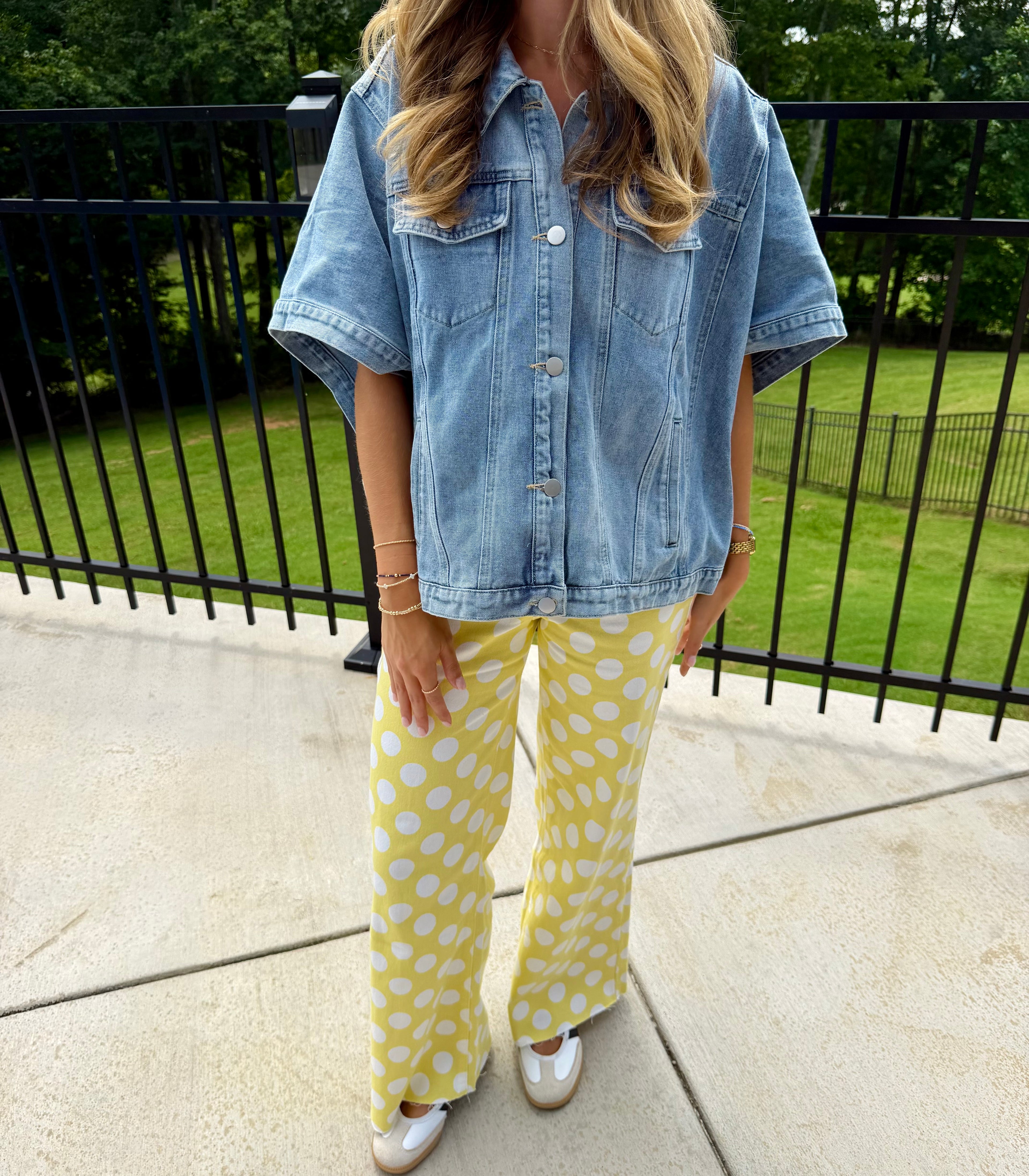 A woman wearing a light blue short-sleeve denim shirt and yellow pants with white polka dots, standing outdoors on a concrete patio in front of a black metal fence and green grass.