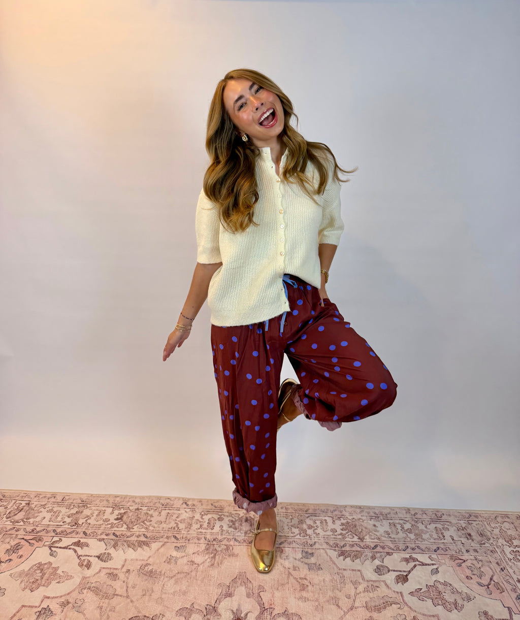 A woman posing in a cream short-sleeve knit top and burgundy pants with blue polka dots, standing on a patterned rug against a plain light gray wall.