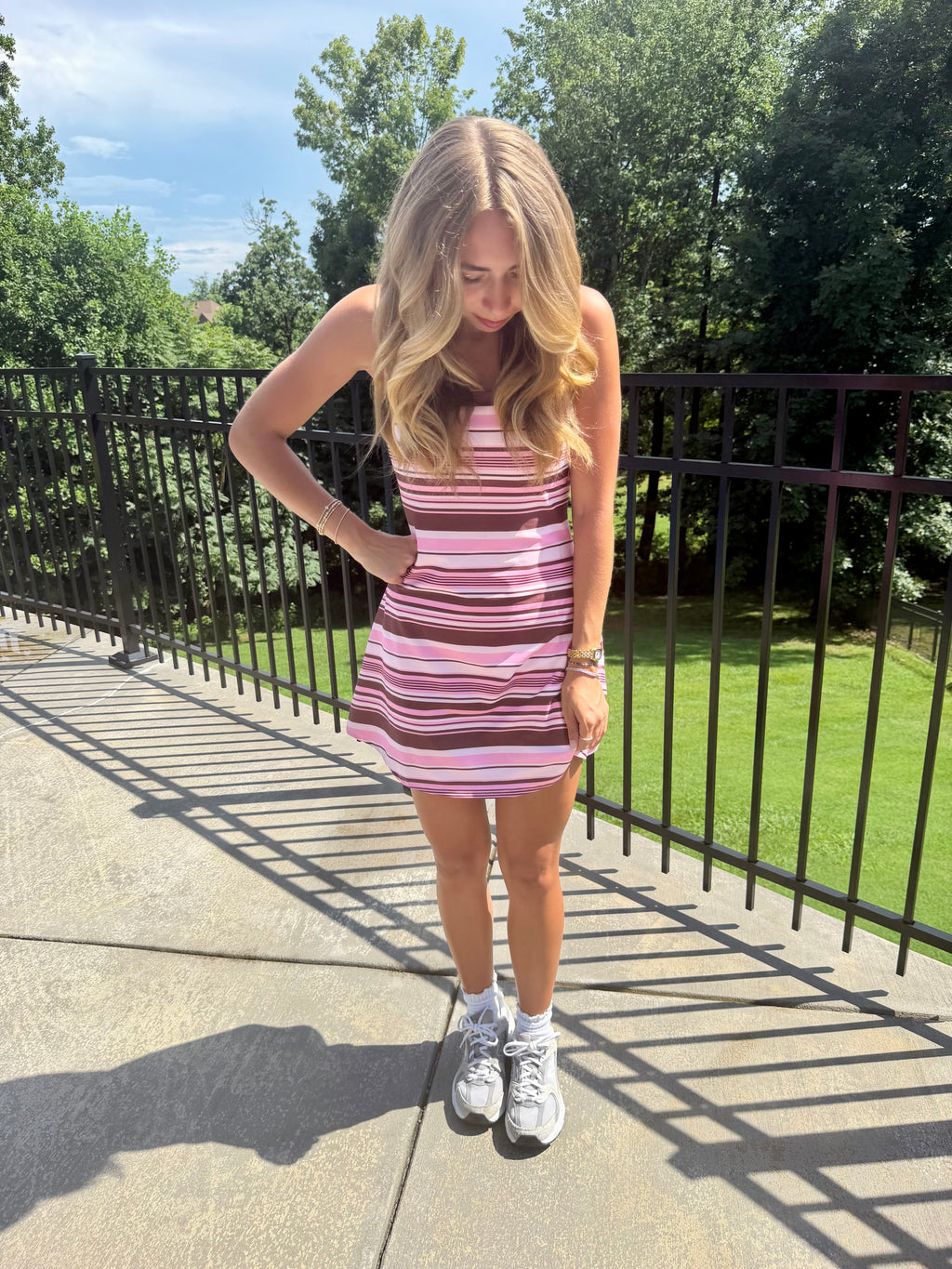 A young woman stands outdoors on a concrete walkway wearing a pink and brown horizontally striped mini dress, white socks, and white sneakers. A black metal fence and green trees are visible in the background.