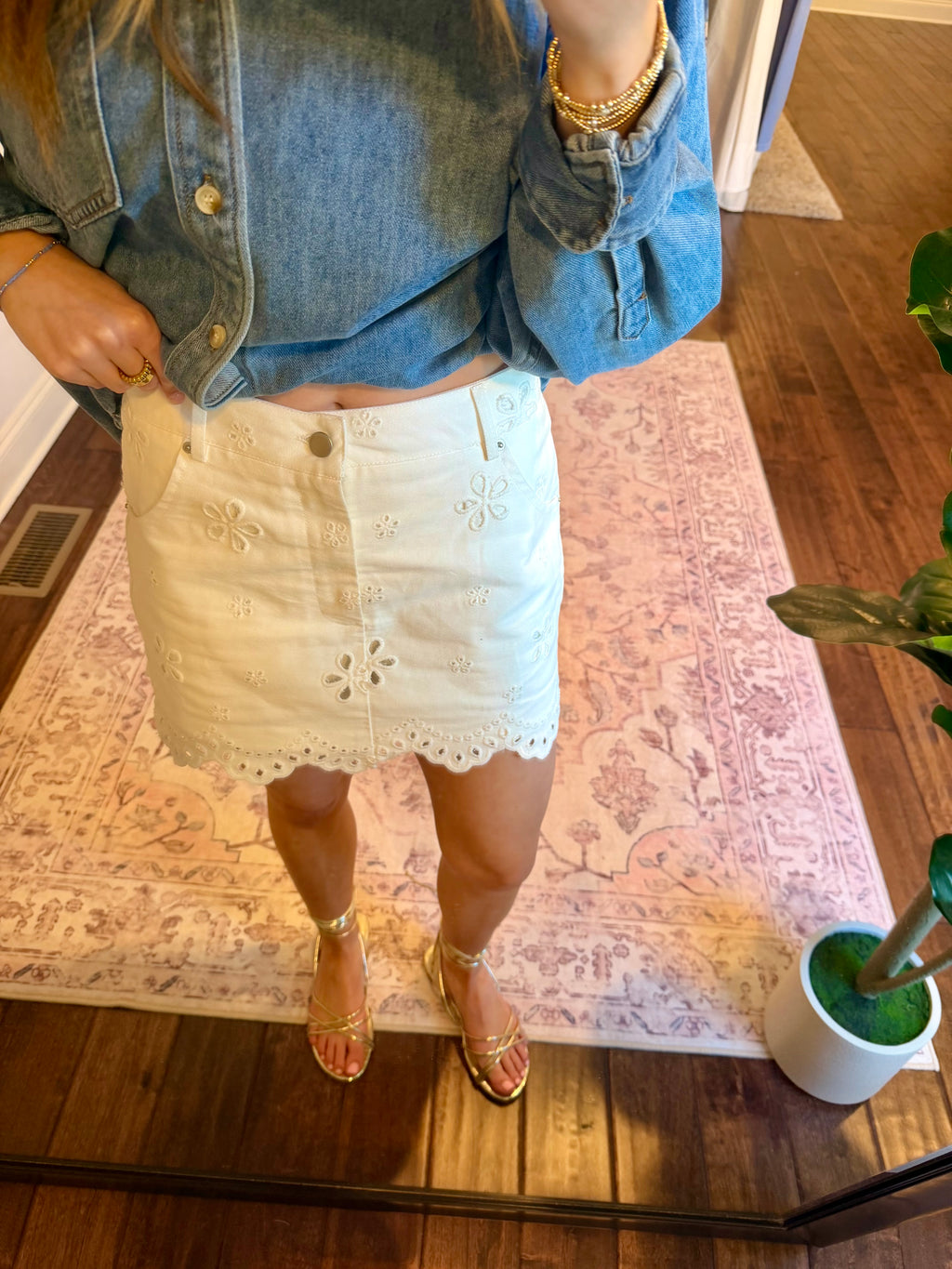 Person taking a mirror selfie wearing a denim jacket and white lace skirt in a room with a rug and plant.