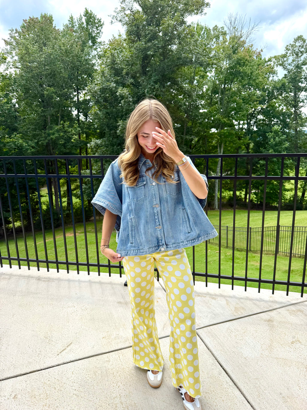 A woman stands outdoors on a concrete patio wearing a light blue short-sleeve denim shirt and yellow pants with white polka dots, with a black metal fence and green trees in the background.