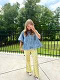 A woman stands outdoors on a concrete patio wearing a light blue short-sleeve denim shirt and yellow pants with white polka dots, with a black metal fence and green trees in the background.