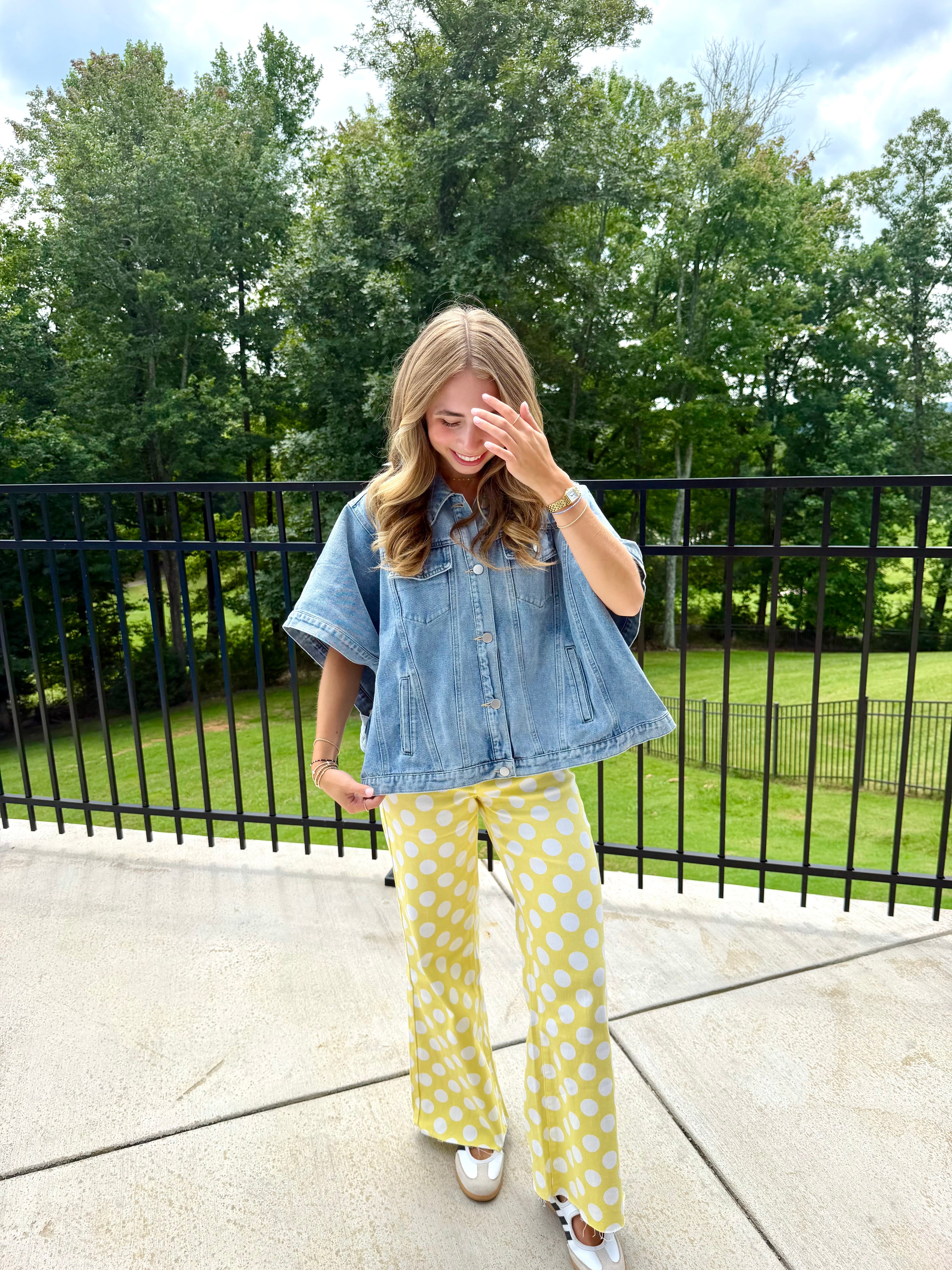 A woman stands outdoors on a concrete patio wearing a light blue short-sleeve denim shirt and yellow pants with white polka dots, with a black metal fence and green trees in the background.