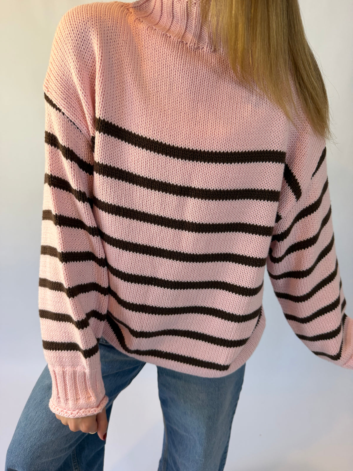 A young woman modeling a pink and brown striped knit sweater, paired with wide-leg blue jeans and pink flat shoes, standing against a light gray studio background.