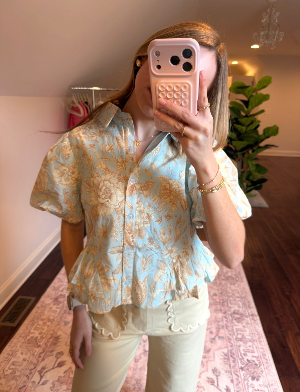 Person wearing a floral blouse taking a mirror selfie in a room with a plant and chandelier.