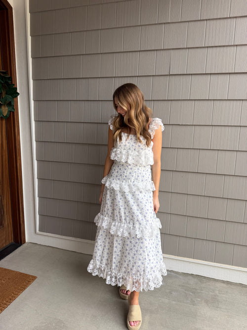 Woman wearing a white dress with ruffles standing against a gray paneled wall.