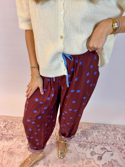 A person wearing burgundy harem-style pants with blue polka dots and a cream cardigan, standing indoors on a patterned rug against a pale pink wall.