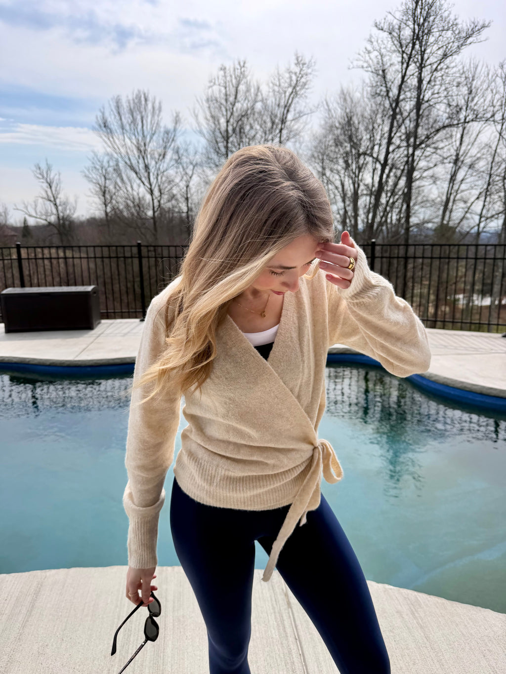 Woman in a beige cardigan standing by a pool with trees and a cloudy sky in the background