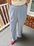 A person wearing blue and white gingham wide-leg pants with ruffled trim at the hem, paired with a cream top and red shoes, standing on a porch with a brick wall and white railing in the background.