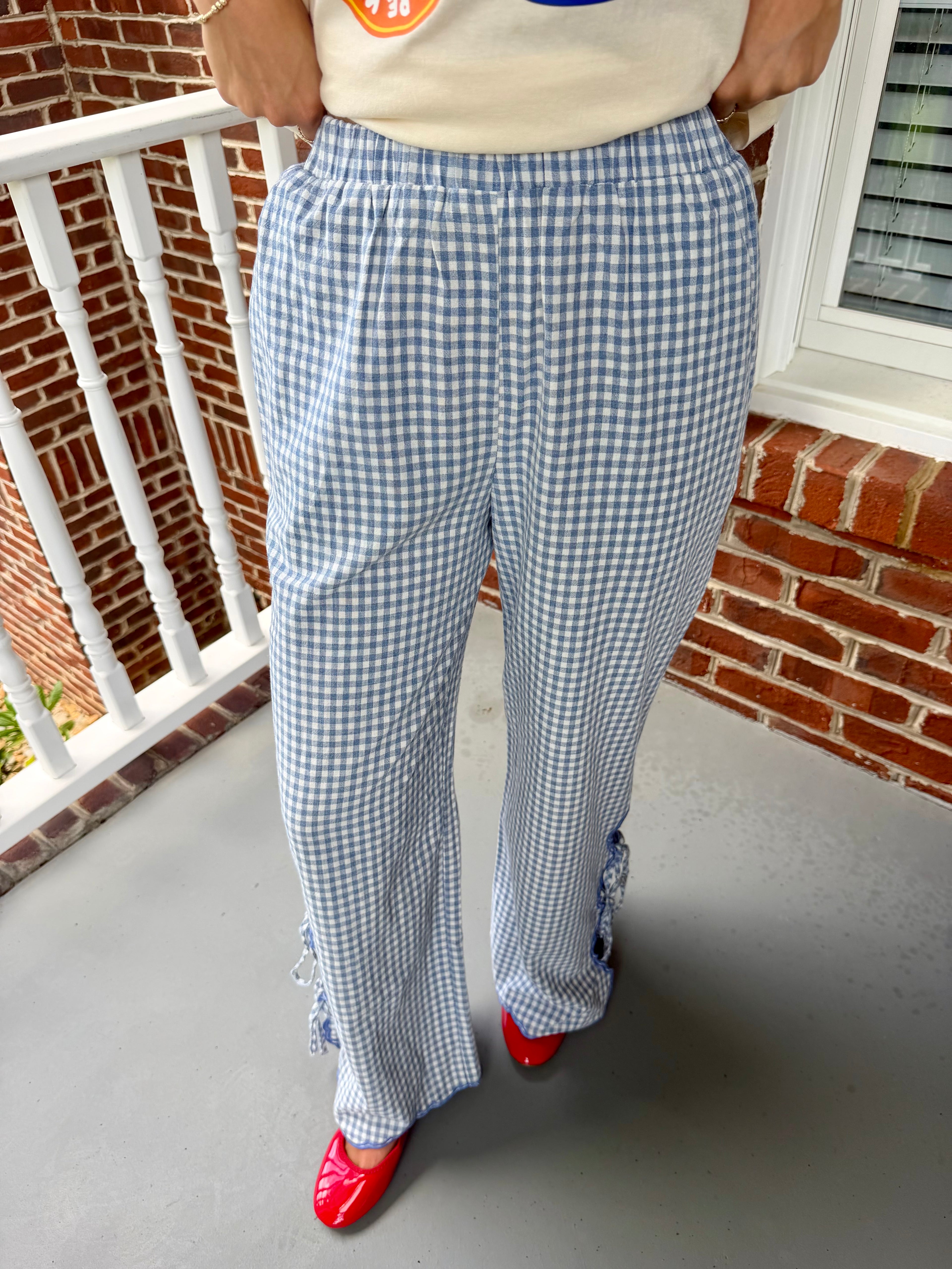 A person wearing blue and white gingham wide-leg pants with ruffled trim at the hem, paired with a cream top and red shoes, standing on a porch with a brick wall and white railing in the background.