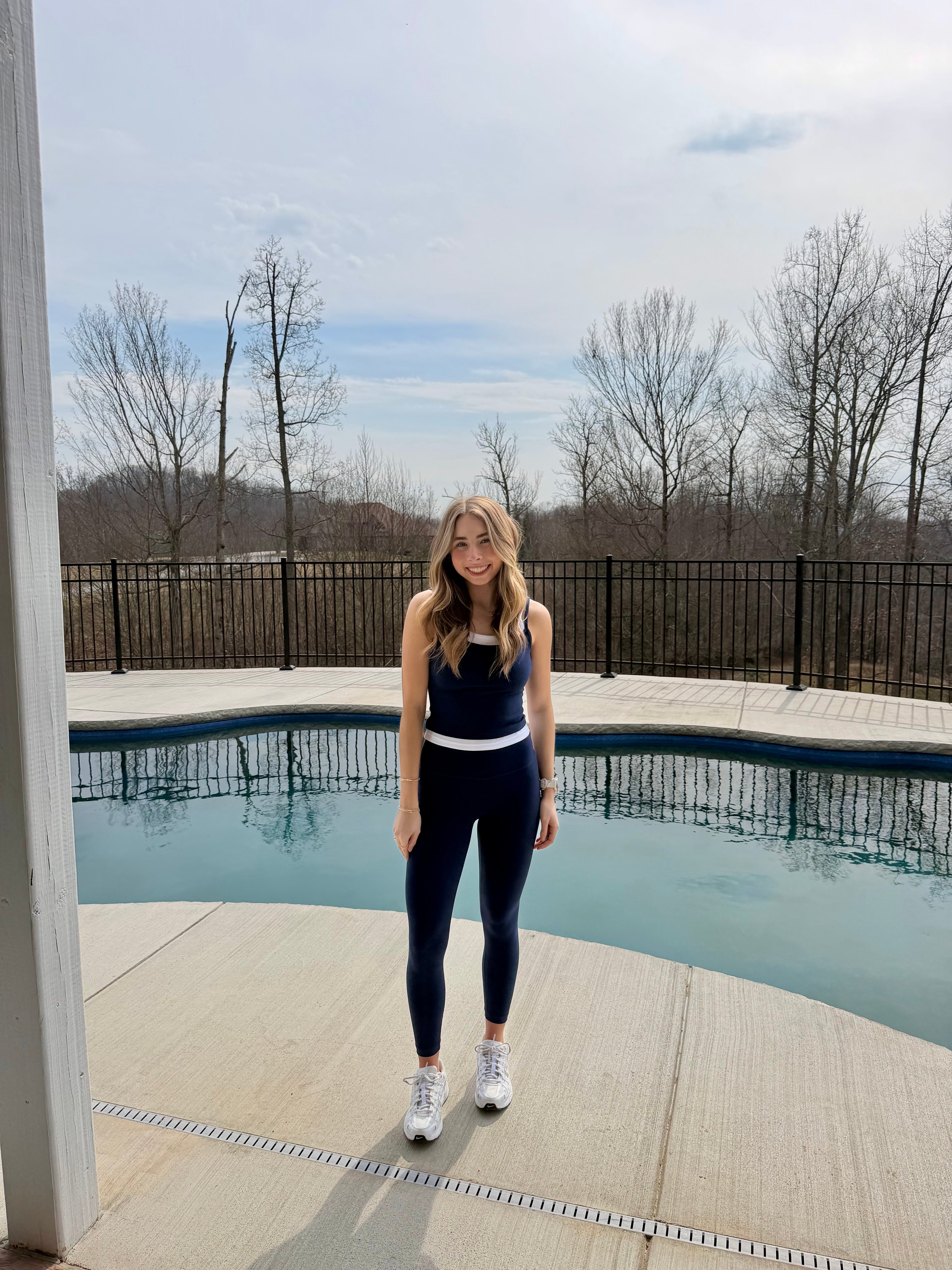 Woman in athletic wear standing by a pool with trees and sky in the background