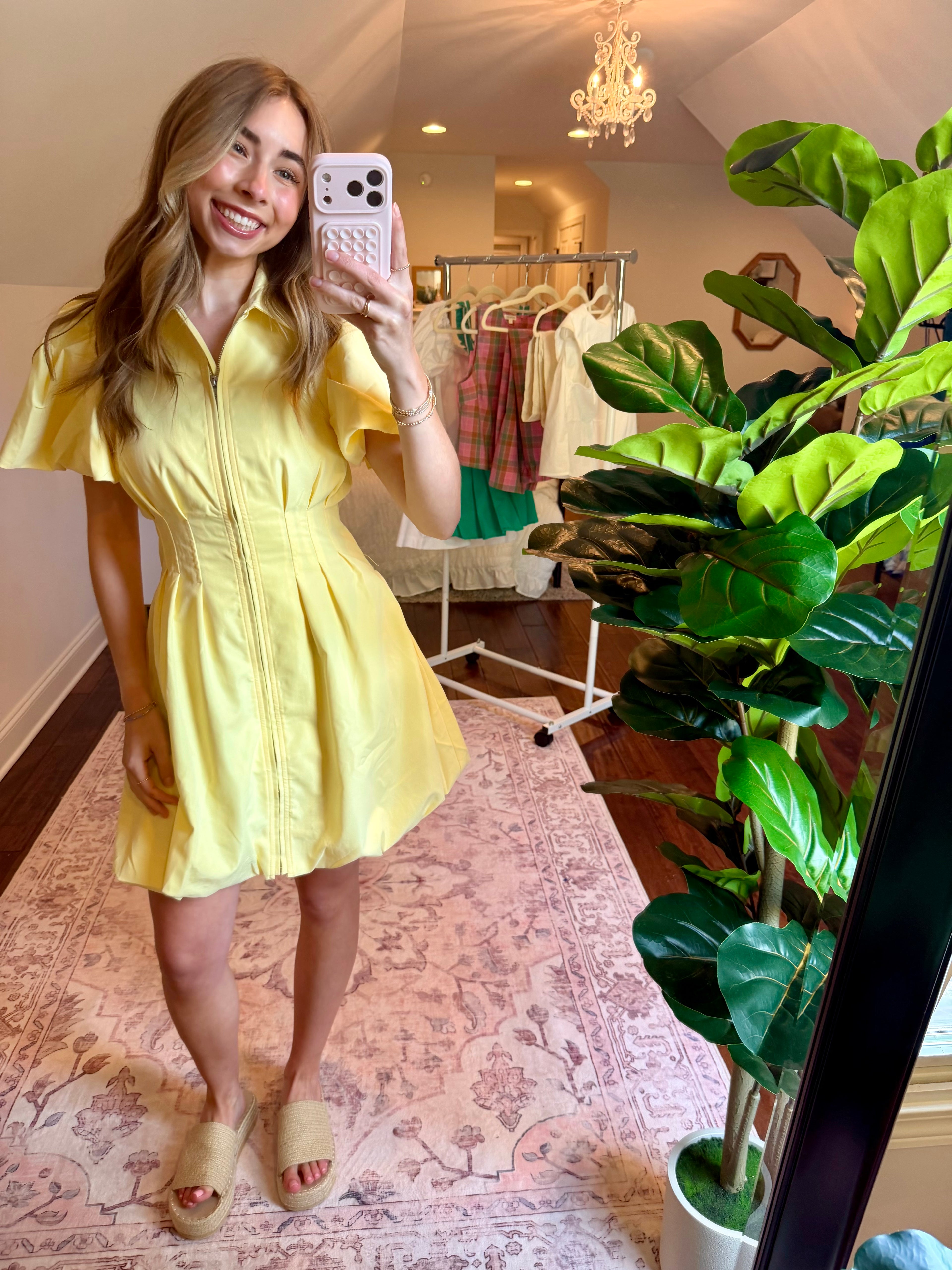 Woman taking a mirror selfie wearing a yellow dress in a room with plants and a chandelier.