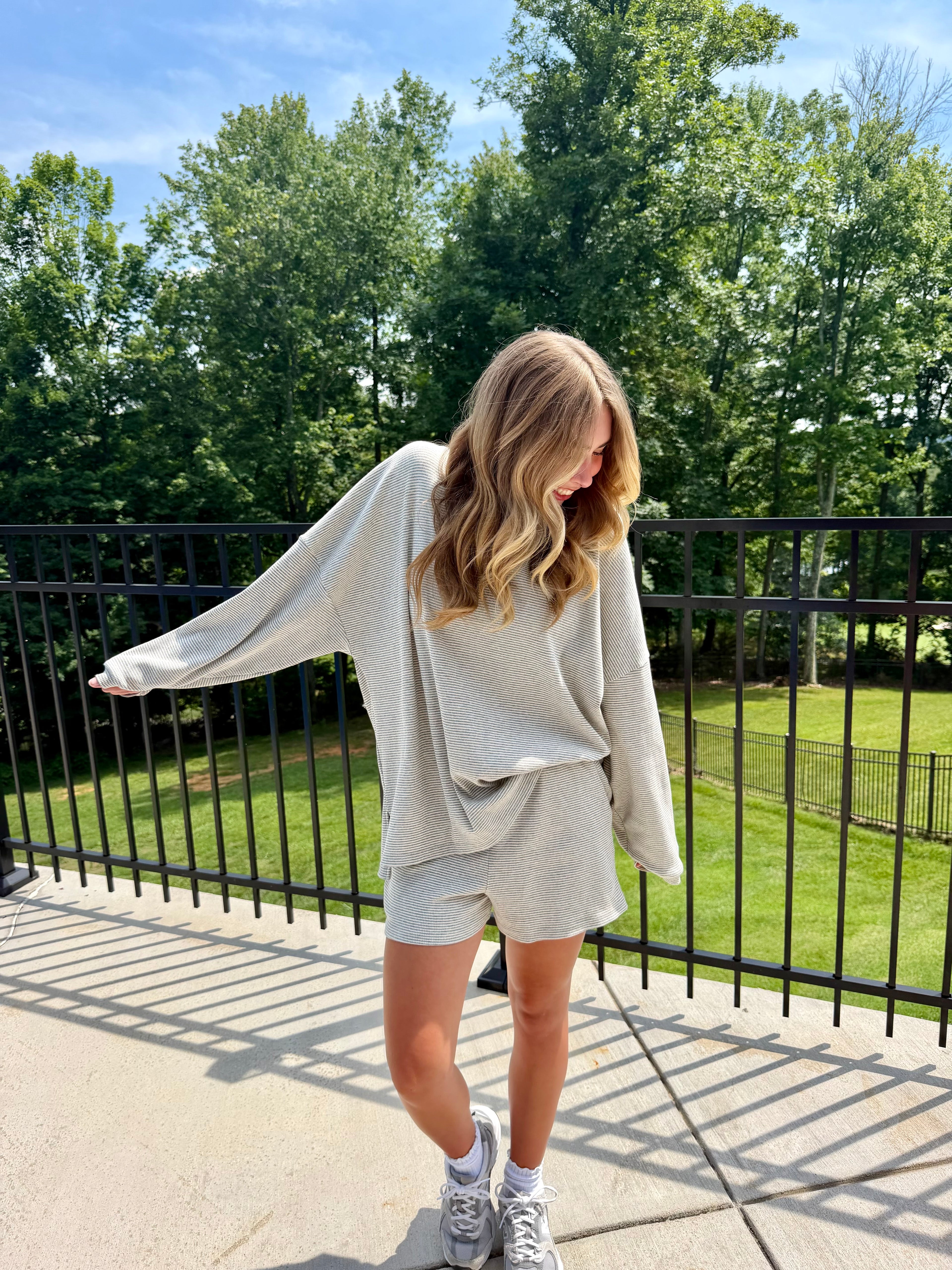 Woman in a light gray outfit standing on a sidewalk with trees in the background