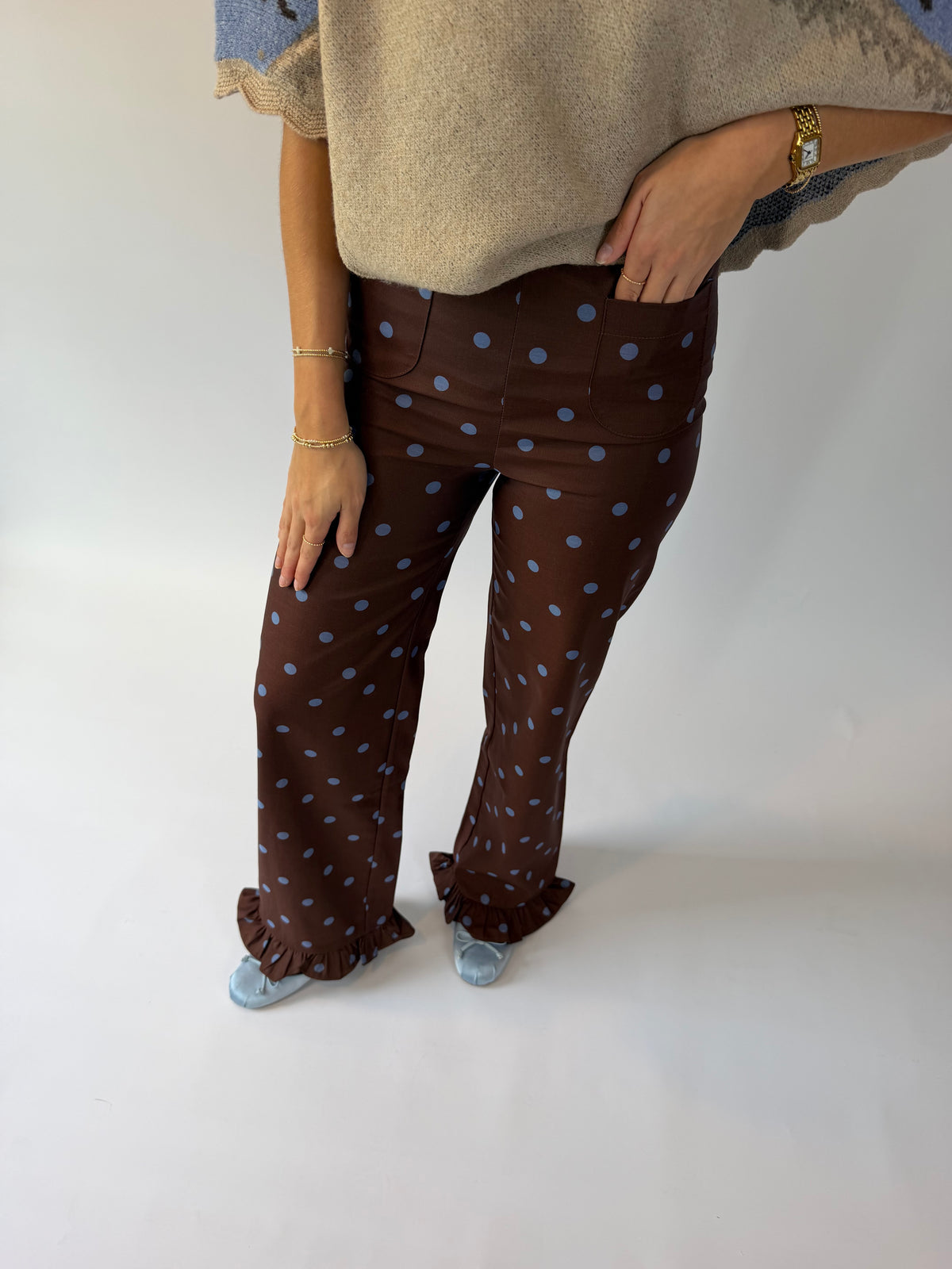 A person wearing brown wide-leg pants with light blue polka dots and ruffled hems, paired with a beige knit top and light blue slip-on shoes, photographed against a plain white studio background.