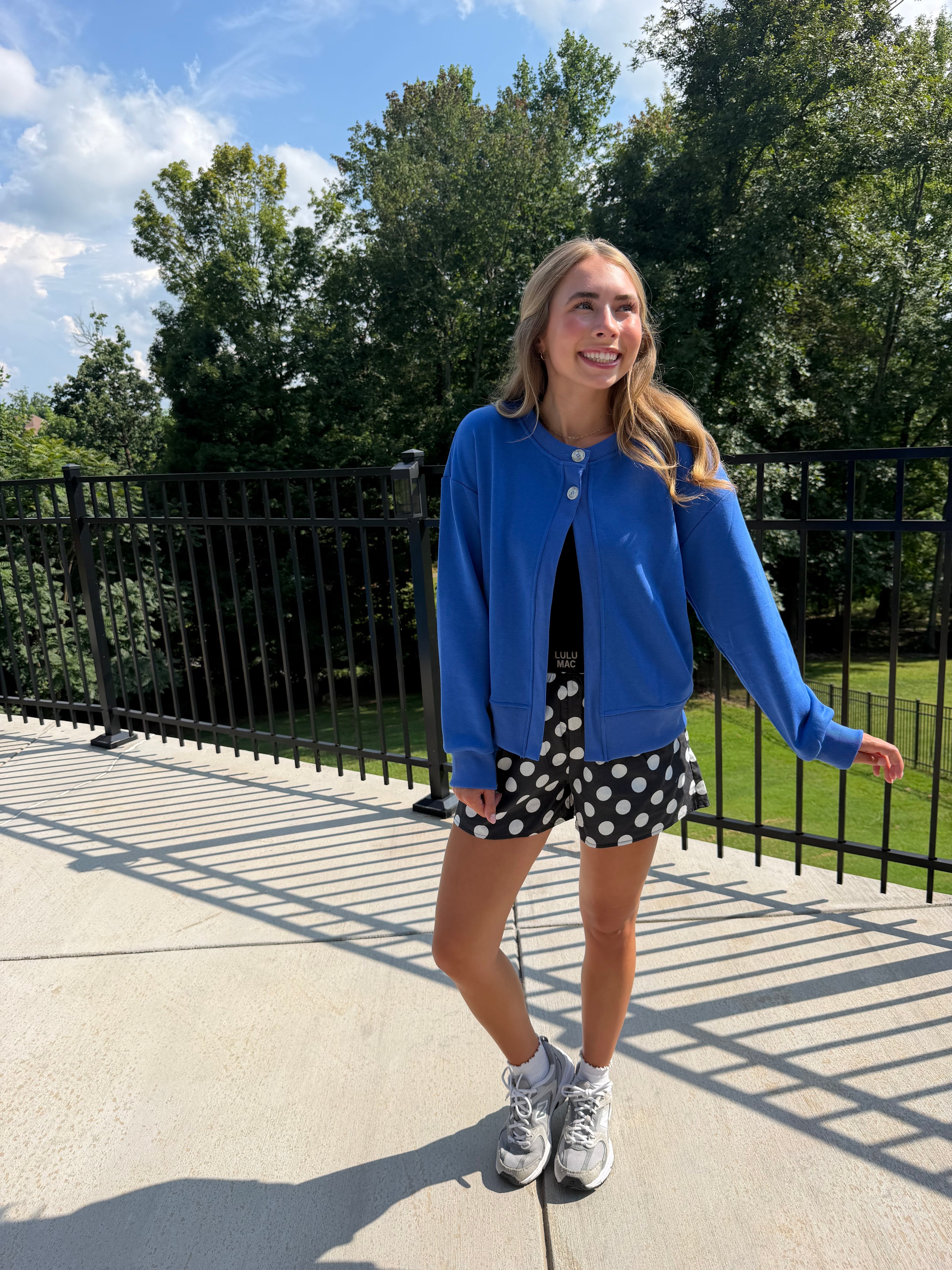 A woman stands outdoors on a concrete walkway wearing a bright blue button-up cardigan, black and white polka dot shorts, white socks, and gray sneakers, with trees and a black fence in the background.