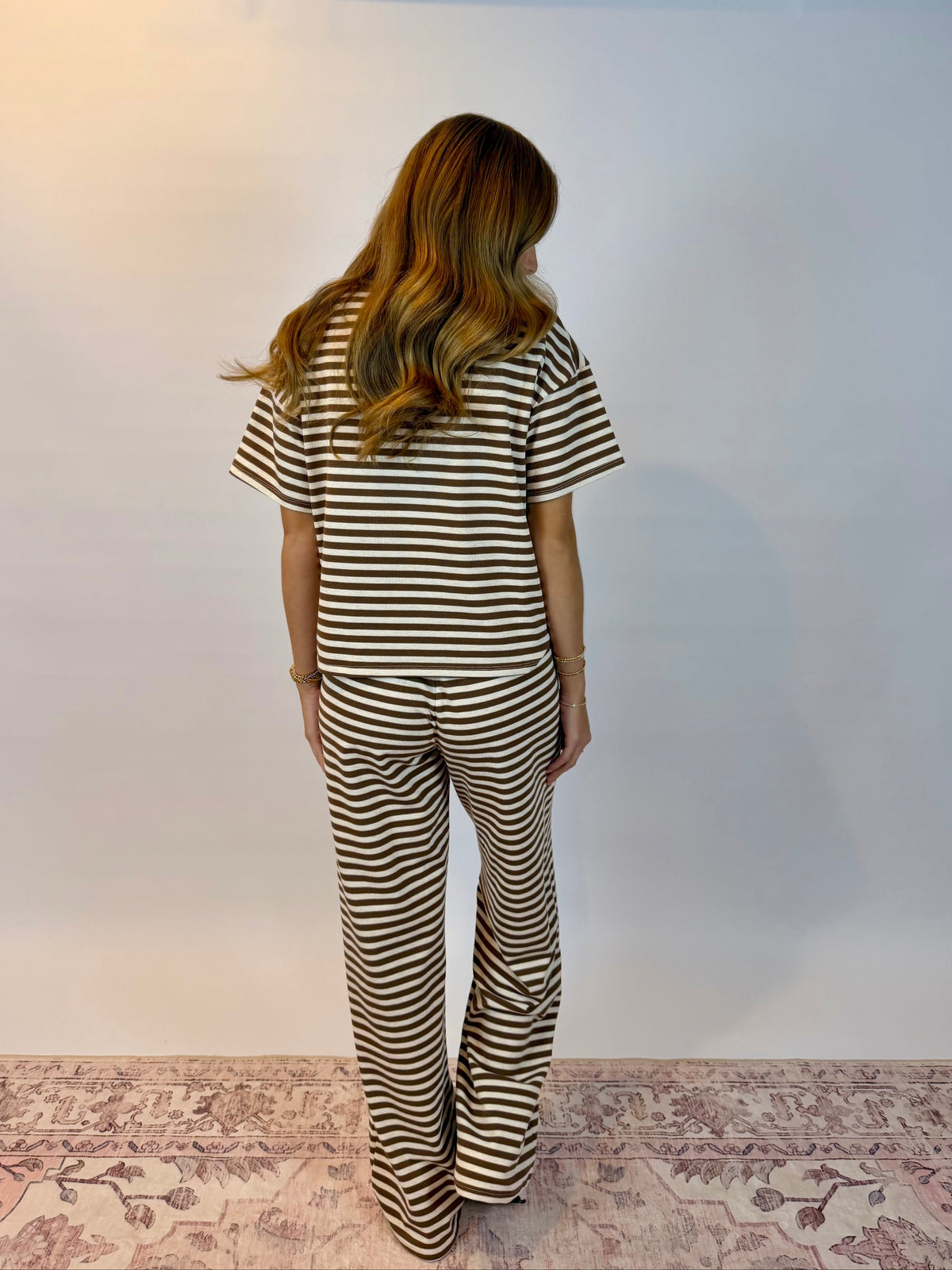 A woman viewed from behind wearing a matching two-piece set consisting of a short-sleeve top and wide-leg pants, both featuring a brown and beige horizontal stripe pattern. She is standing on a patterned rug against a plain white studio backdrop.