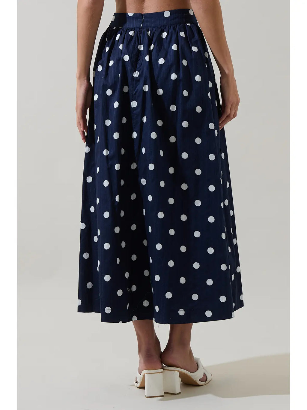 A navy blue midi skirt with a white polka dot pattern, shown on a model from the waist down wearing white block-heel slide sandals against a light gray studio background.