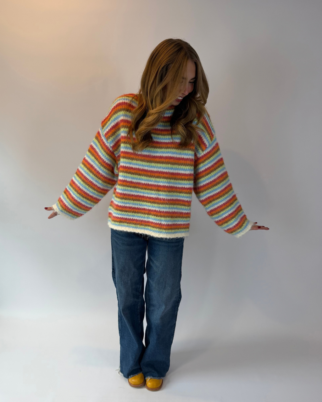 A woman wearing a fuzzy, oversized striped sweater with blue jeans and yellow shoes, standing against a plain light gray studio background.